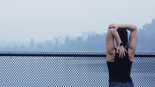 Athletic brunette woman stretches her triceps nxt to a dark chain-link fence near a lake in the rain