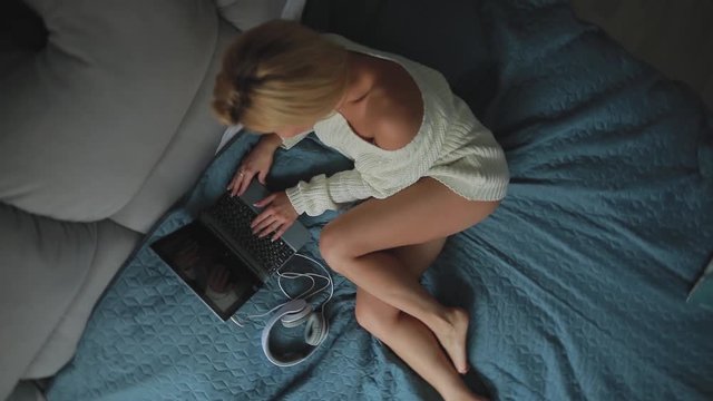Sexy woman using laptop at home lying on a cozy bed in apartment room in sweater and panties, top view, rotating camera