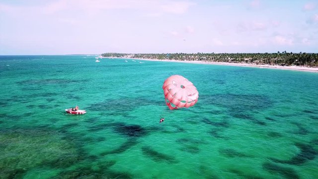 Aerial, orbit, drone shot, around a boat and people paragliding, with a red para sail over turquoise sea, just outside a paradise beach, on a sunny, summer day, in Dominican republic