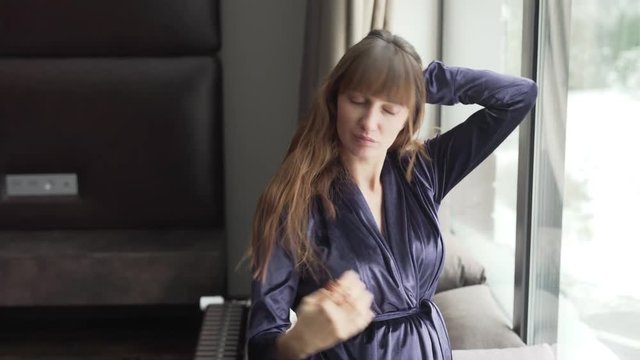 A young pretty brunette with ponytail hair dressed in dark blue gown, making her hair loose while looking in the mirror. She's sitting near sunny window. Businesslady concept