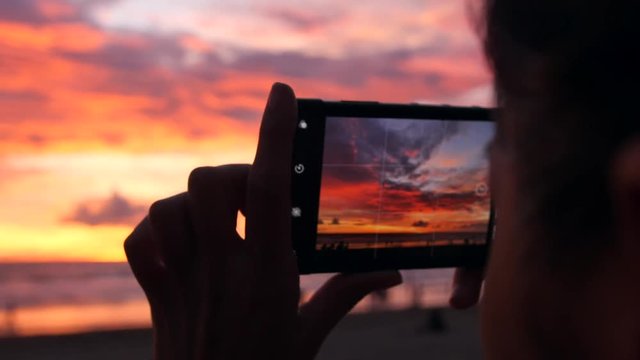 Young Tourist Woman Taking Photos with Mobile Phone Camera of Amazing Sunset at the Beach. Female Hands Holding Smartphone and Doing Nature Photography. 4K Slowmotion. Bali, Indonesia.