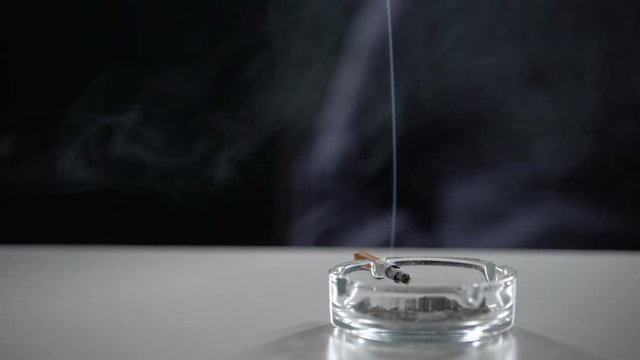 Cigarette burning in ash tray isolated on dark background, nicotine addiction