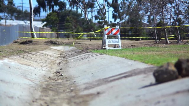 An empty desolate park enclosed by a chain link fence where the wind blows caution tape attached to a construction barrier just off a cement path.