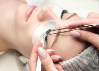 Obraz na płótnie Canvas Eyelash extension procedure. Woman face and experts fingers with tweezers, close up, selective focus