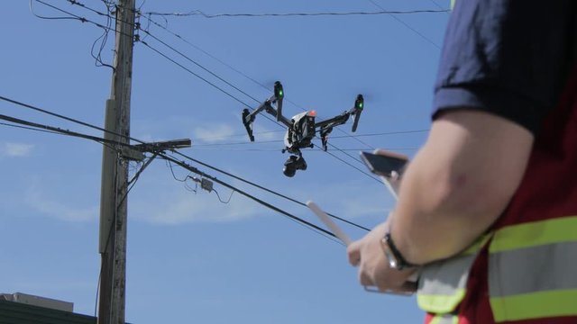 A drone is hovering in front of telephone lines while being operated by a worker in a safety vest with a remote control. Drone is in focus while worker and remote are slightly blurry.