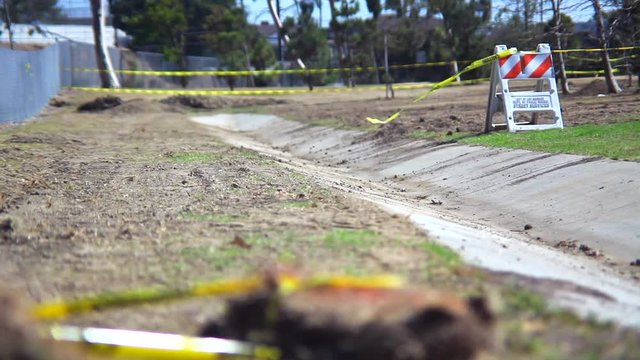 An empty desolate park enclosed by a chain link fence where the wind blows caution tape attached to a construction barrier just off a cement path.