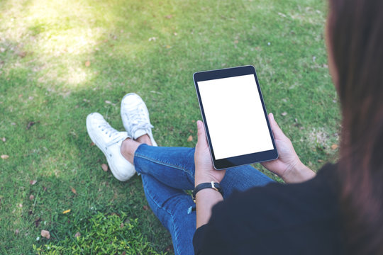 Mockup image of a woman holding and using black tablet pc with blank white desktop screen while sitting in the outdoors