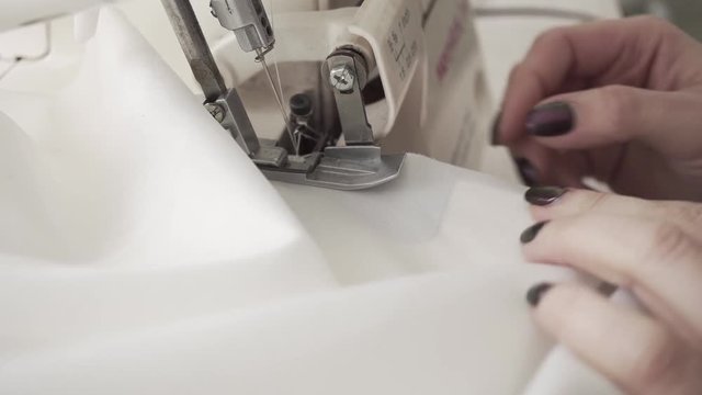 Seamstress sews clothes made of white cloth on a sewing machine. Work by the light of the built-in hardware lamp. Steel needle with looper and presser foot close-up