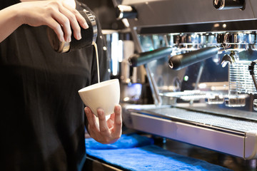 Closeup image of female barista pouring milk and preparing fresh cappuccino while standing in front of the coffee machine at coffee shop in morning