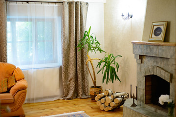 fireplace with firewood in the room for rest in the village house. next to a green plant and a sofa, on the floor a carpet - obrazy, fototapety, plakaty