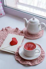 Cup of scented pink tea on crocheting napkin, teapot and envelope on windowsill. Valentines day. Romance Good morning