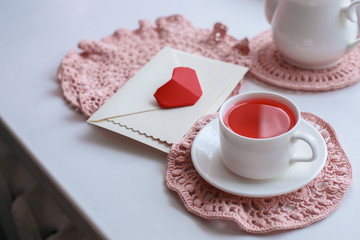 Cup of scented pink tea on crocheting napkin, teapot and envelope on windowsill. Valentines day. Romance Good morning