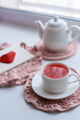 Cup of scented pink tea on crocheting napkin, teapot and envelope on windowsill. Valentines day. Romance Good morning
