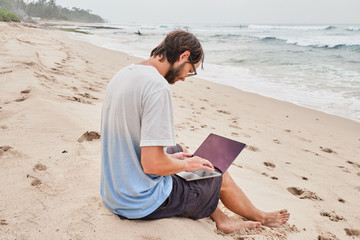 A young man works at the laptop on the beach. Freelancer working on the beach by the ocean.
