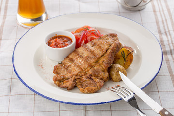 Grilled pork steak with potato wedges, tomatoes and ketchup on white plate on textile background