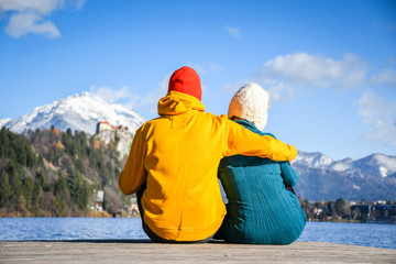 Fototapeta premium Couple in love hugging together with colorful cloths sitting and relaxing on a wooden pier on a clear sky sunny winter day view from the back, Bled