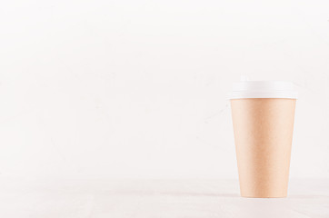 Coffee packing mockup -  kraft brown paper cup with white cap on white wood board with copy space, coffee shop interior. Modern elegant concept for branding identity, advertising, design.