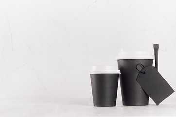 Coffee mockup - different size black paper cups with white caps, blank label and sugar bag on white wood table with copy space, coffee shop interior. 