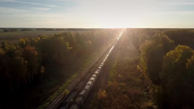 aerial two freight trains with cisterns and high-sided open wagons move in opposite directions through wood at sunset