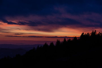 Trees silhouettes against a beautifully colored sky at dusk, with mountains layers in the background