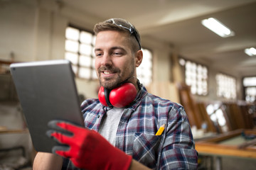 Portrait of middle aged professional worker carpenter with ear protectors using tablet in carpentry...