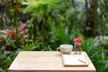 Coffee cup notebook,brown pencil and beautiful red dried flower in glass vase on wood table with fresh green garden background,outdoor workspace