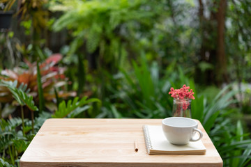 Coffee cup notebook,brown pencil and beautiful red dried flower in glass vase on wood table with fresh green garden background,outdoor workspace