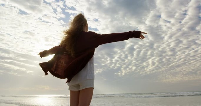 Rear view of young woman with arms outstretched looking at sea on the beach 4k