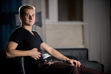 Young attractive smiling guy in dark t-shirt sitting on a couch and looking in the camera