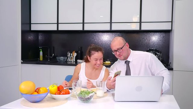 cheerful wife and husband sitting in kitchen in morning, viewing funny pictures in smartphone