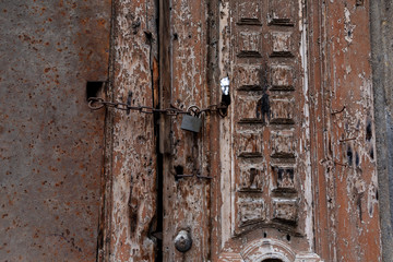 Old wooden door with flaky brown paint and carved pattern. Closed on a modern lock on a rusty chain. Wooden and metal old collapsing surface