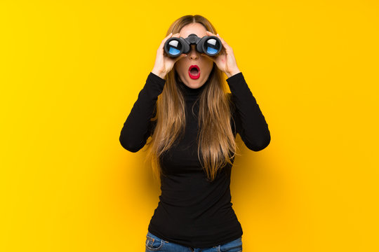 Young pretty woman over yellow background and looking in the distance with binoculars