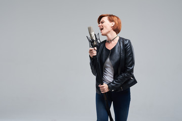 Portrait of rock singer wearing leather jacket and keeping static mic, sings a song loudly on grey...