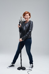 Full-length portrait of rock singer wearing leather jacket and keeping static mic, sings a song loudly on grey background. Concept of rock music and rave