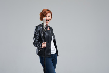 Portrait of rock singer wearing leather jacket and keeping static mic, sings a song loudly on grey...