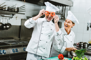 smiling female cook looking at male chef holding pepper slices in front of face in restaurant...