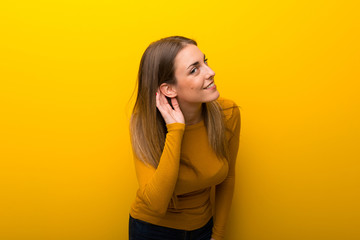 Young woman on yellow background listening to something by putting hand on the ear