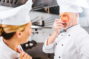 female cook looking at male chef holding pepper slice in front of face in restaurant kitchen