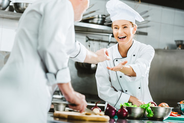 smiling female chef in uniform taking ingredient from male colleague during cooking in restaurant kitchen