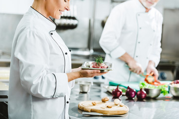 cropped view of female chef in uniform holding meat dish on plate with colleague cooking on background in restaurant kitchen