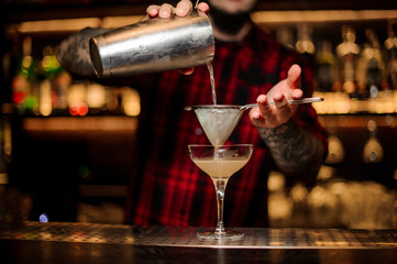 Barman pouring juicy alcoholic cocktail from shaker into cocktail glass using strainer