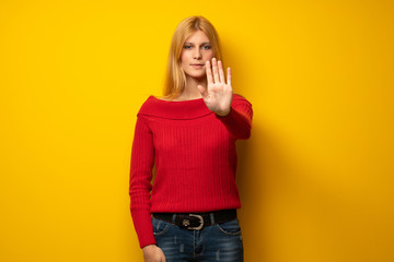 Blonde woman over yellow wall making stop gesture denying a situation that thinks wrong