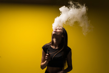 Young brunette woman standing and vaping on yellow background