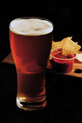 A glass of beer on a black background, nachos chips with sauce in the background.