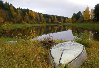 Autumn forest landscape. In the foreground in the grass turning yellow is an inverted fishing boat....