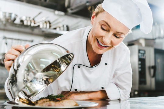 Beautiful Female Chef In Uniform Holding Dome From Serving Tray With Meat Dish In Restaurant Kitchen