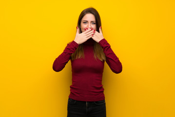 Woman with turtleneck over yellow wall covering mouth with hands for saying something inappropriate