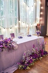 Wedding presidium in restaurant, copy space. Banquet table for newlyweds with flowers, monstera leaves, violet cloth and bulbs. Lush floral arrangement. Luxury wedding decorations