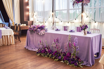 Wedding presidium in restaurant, copy space. Banquet table for newlyweds with flowers, monstera leaves, violet cloth and bulbs. Lush floral arrangement. Luxury wedding decorations