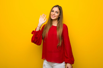 Young girl with red dress over yellow wall saluting with hand with happy expression